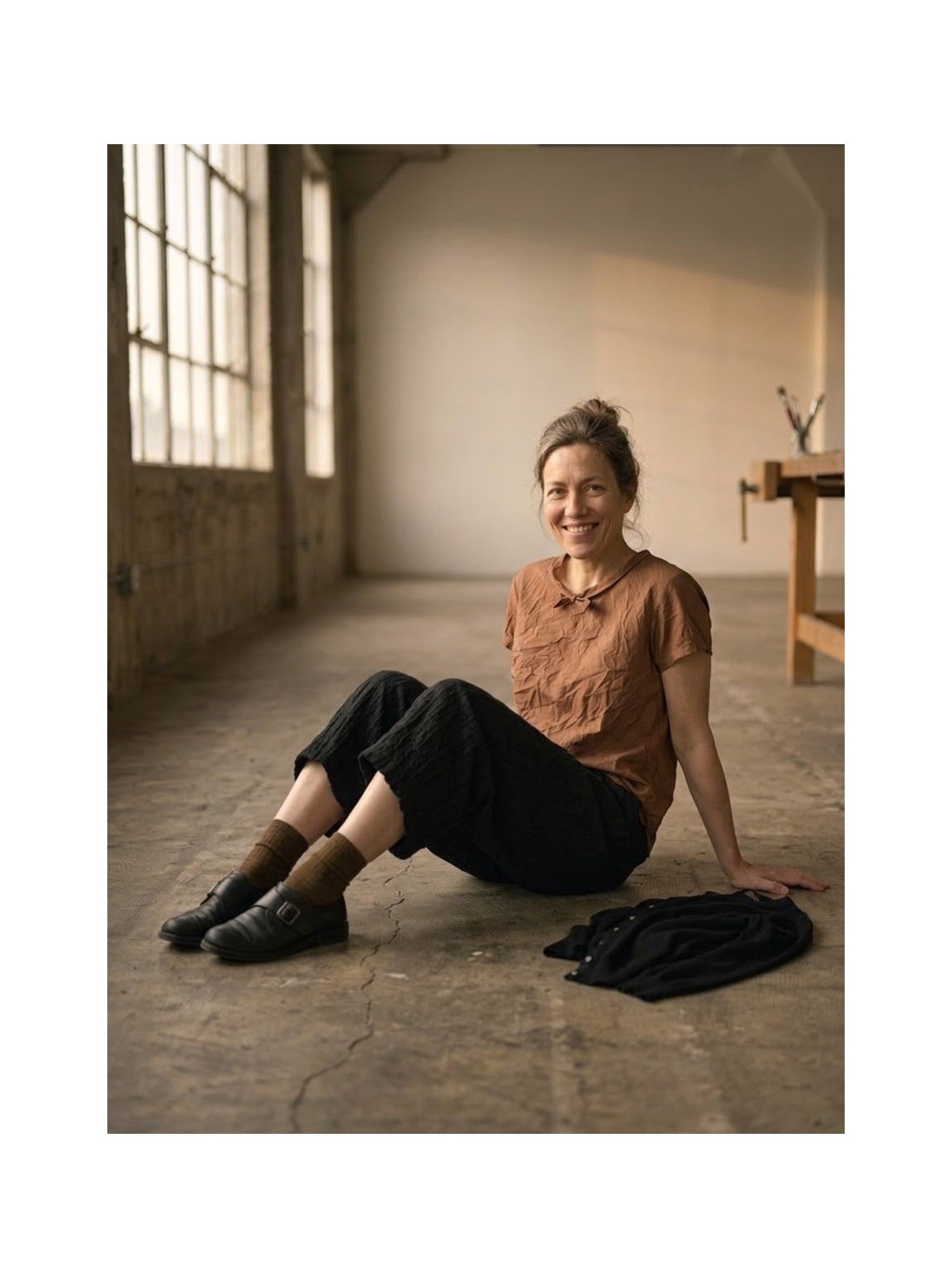 A woman sits on the concrete floor of a rustic studio, wearing a rust-brown short-sleeve button-up shirt, black cropped trousers, and dark brown loafers. A matching rust-brown shirt and black trousers are laid out on the floor beside her.